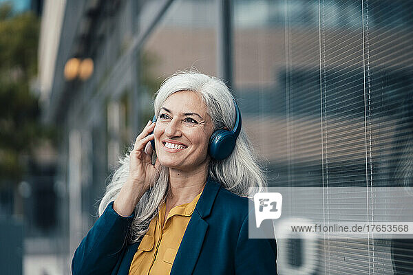 Happy businesswoman listening music through wireless headphones by glass wall