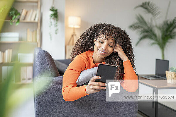 Smiling woman with curly hair using mobile phone on sofa at home