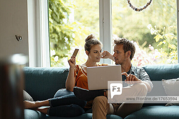 Woman holding credit card sitting by boyfriend with laptop on sofa in living room