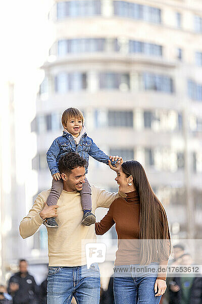 Happy man carrying son on shoulder walking with young woman in front of building
