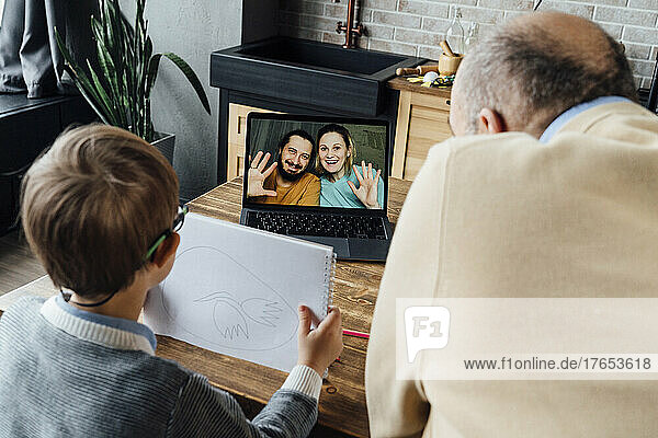Boy showing drawing to mother and father through video call on laptop sitting by grandfather at home