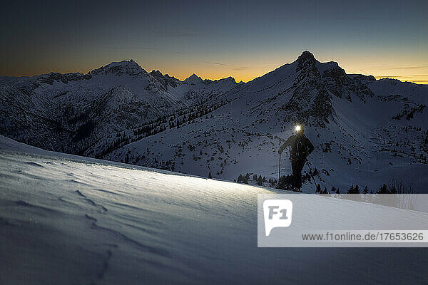 Mann mit Stirnlampe geht bei Sonnenuntergang auf verschneiten Berg
