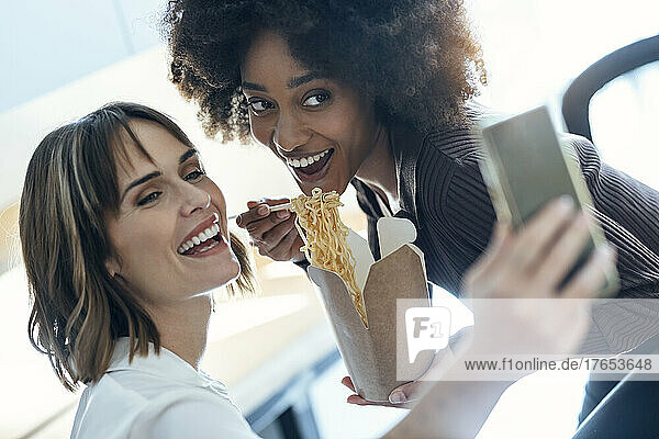 Happy businesswoman taking selfie with colleague having noodles in office