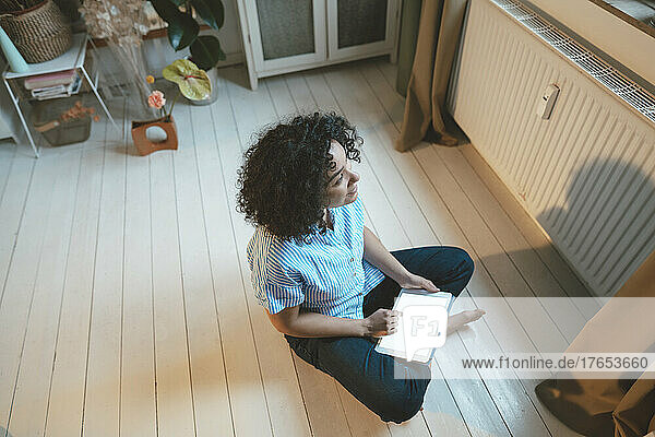 Thoughtful woman with tablet PC sitting on floorboard at home