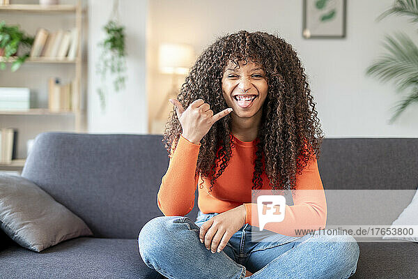 Young woman gesturing shaka sign sitting on sofa at home