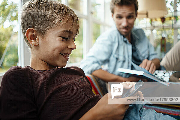 Glücklicher blonder Junge  der zu Hause beim Vater sitzt und sein Smartphone benutzt