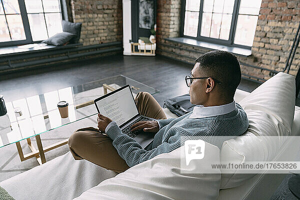 Young freelancer wearing eyeglasses using laptop in living room at home