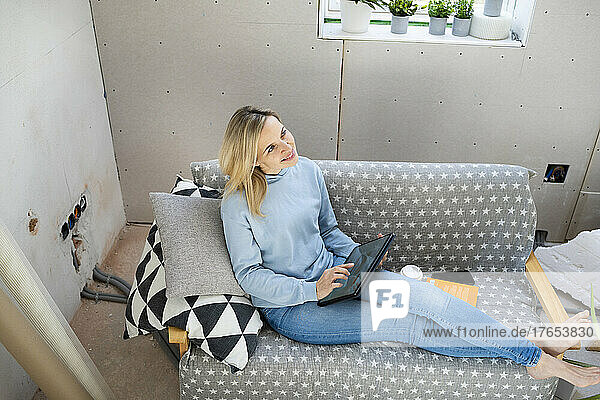 Happy blond woman with tablet PC sitting on sofa in attic
