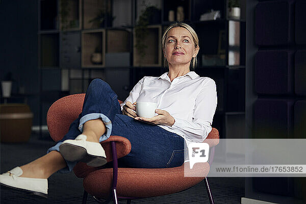 Geschäftsfrau hält Kaffeetasse in der Hand und sitzt mit gekreuzten Beinen auf einem Stuhl