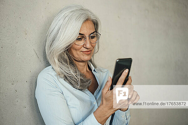 Businesswoman wearing eyeglasses using mobile phone in front of wall