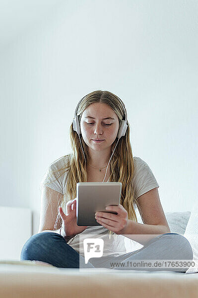 Woman using tablet PC listening music through headphones sitting on bed