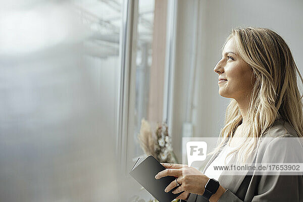 Smiling businesswoman holding diary looking through window in office