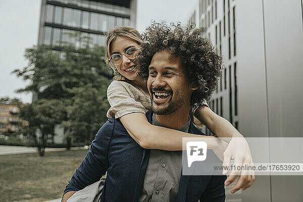 Happy young man piggybacking girlfriend in front of modern building
