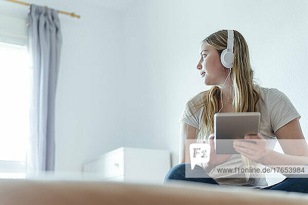 Woman listening music through headphones sitting with tablet PC on bed at home