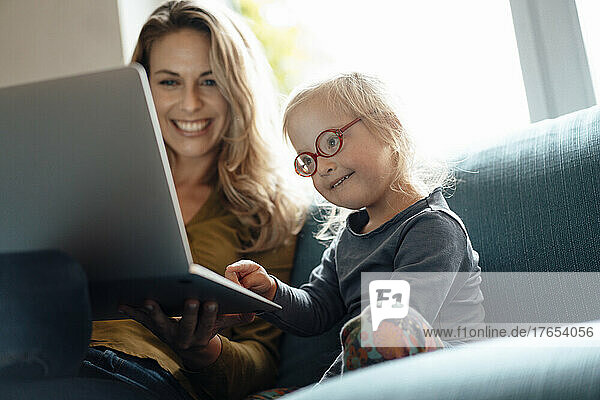 Little girl wearing eyeglasses using laptop sitting by mother on sofa in living room