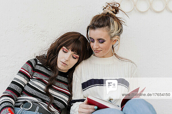 Woman leaning on friend sitting with book in front of white wall