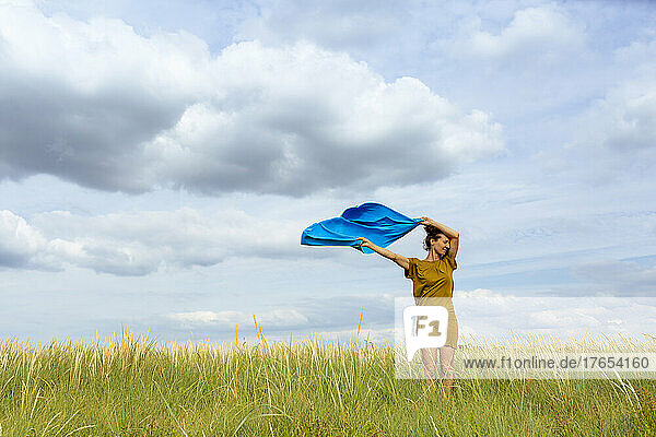 Woman holding aloft wind-swept blue scarf at meadow