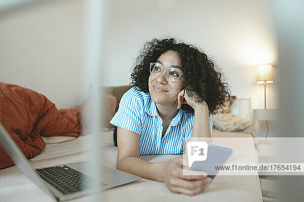 Thoughtful woman with laptop and mobile phone lying on bed at home