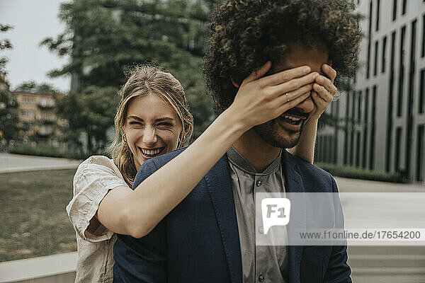 Happy young woman covering boyfriend's eyes with hand in front of modern building