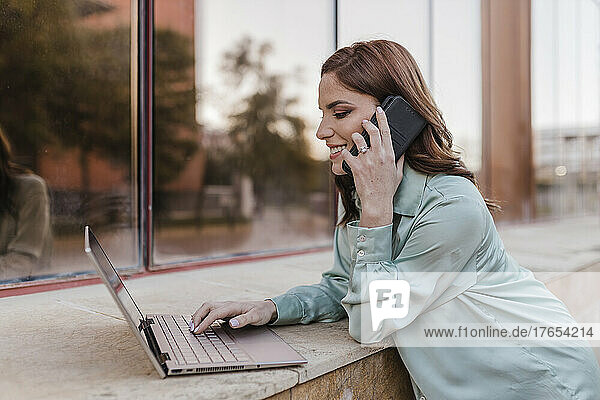 Smiling businesswoman using laptop and talking on smart phone