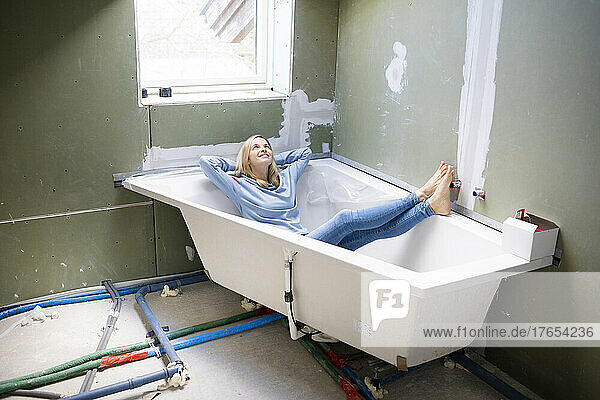 Glückliche blonde Frau mit den Händen hinter dem Kopf sitzt in der Badewanne auf dem Dachboden