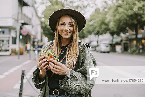 Happy young woman with sandwich standing on street