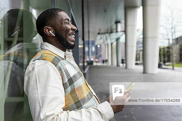 Happy man with eyes closed holding mobile phone leaning on glass wall
