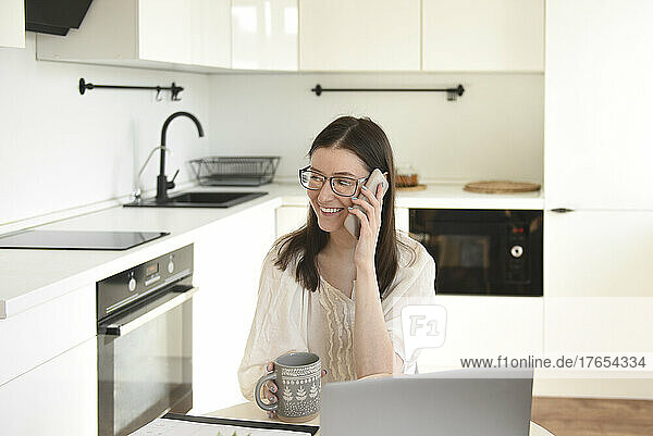 Smiling freelancer talking on mobile phone sitting with laptop in kitchen