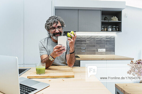 Smiling senior man with apple using smart phone at kitchen island