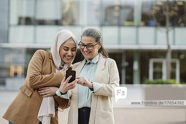 Happy businesswoman wearing eyeglasses sharing smart phone to colleague at office park