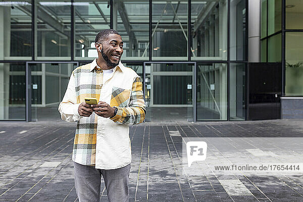 Smiling man holding mobile phone standing in front of glass building