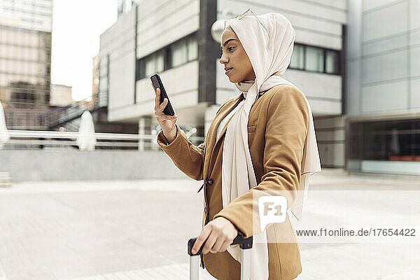 Businesswoman wearing hijab surfing net through smart phone standing outside modern building