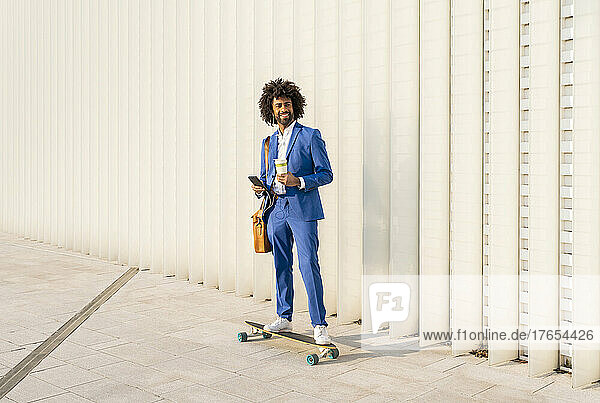Smiling businessman holding smart phone and disposable cup standing on skateboard in front of wall