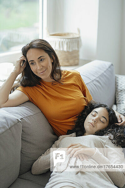 Teenage girl resting in lap of thoughtful mother on sofa at home
