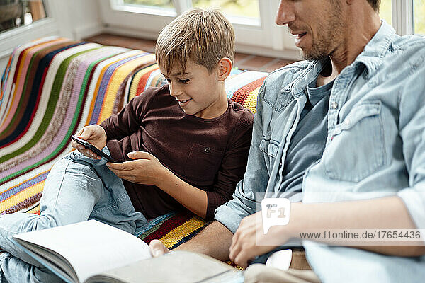 Man with book looking at son using smart phone sitting on sofa ta home