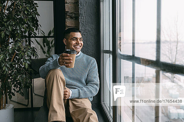 Happy young man with disposable coffee cup looking through window sitting at home