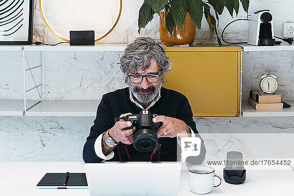 Senior freelancer holding camera sitting with laptop at home