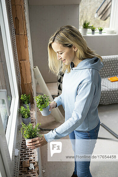 Smiling blond woman decorating attic with plants on window sill