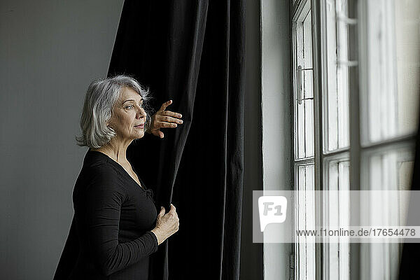 Senior woman standing by curtain looking through window at home