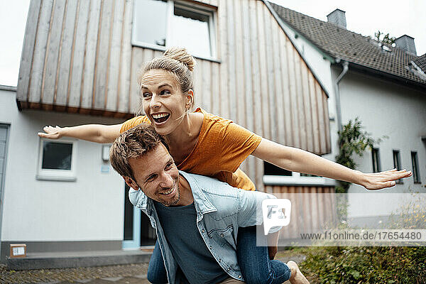 Happy man giving piggyback ride to woman with arms outstretched enjoying at backyard