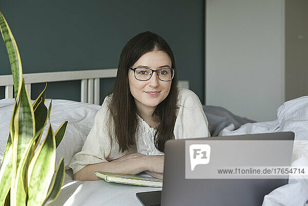 Smiling woman wearing eyeglasses with laptop lying on bed at home