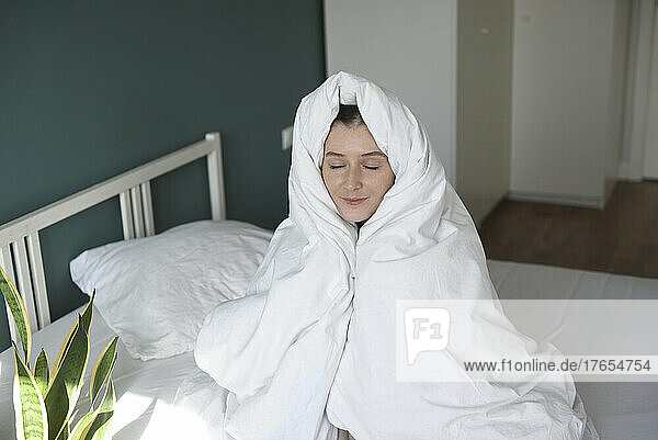 Woman wrapped in blanket sitting on bed at home