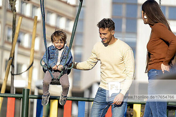 Smiling man pushing son on swing standing by woman in playground