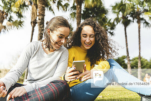 Happy friends sharing mobile phone sitting in park on sunny day