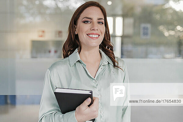 Happy businesswoman with diary standing in front of glass window