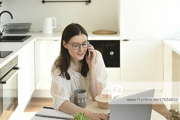 Freelancer using laptop talking on mobile phone sitting at table in kitchen
