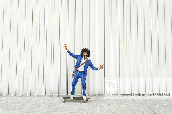 Playful businessman standing with arms outstretched on skateboard in front of wall