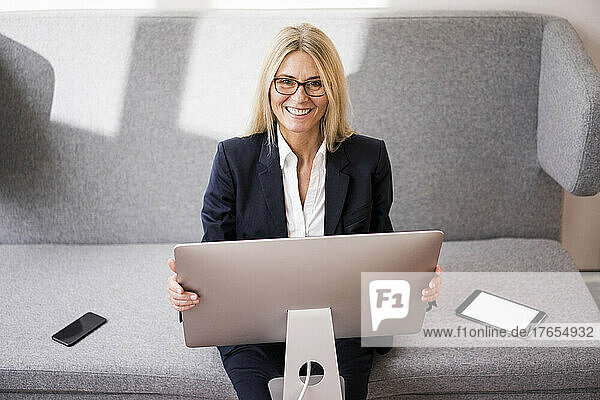 Smiling businesswoman with computer sitting on sofa in office