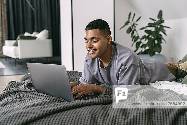 Smiling young man using laptop lying on bed at home