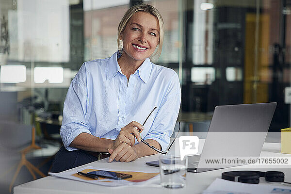Smiling businesswoman holding eyeglasses and laptop at desk in office
